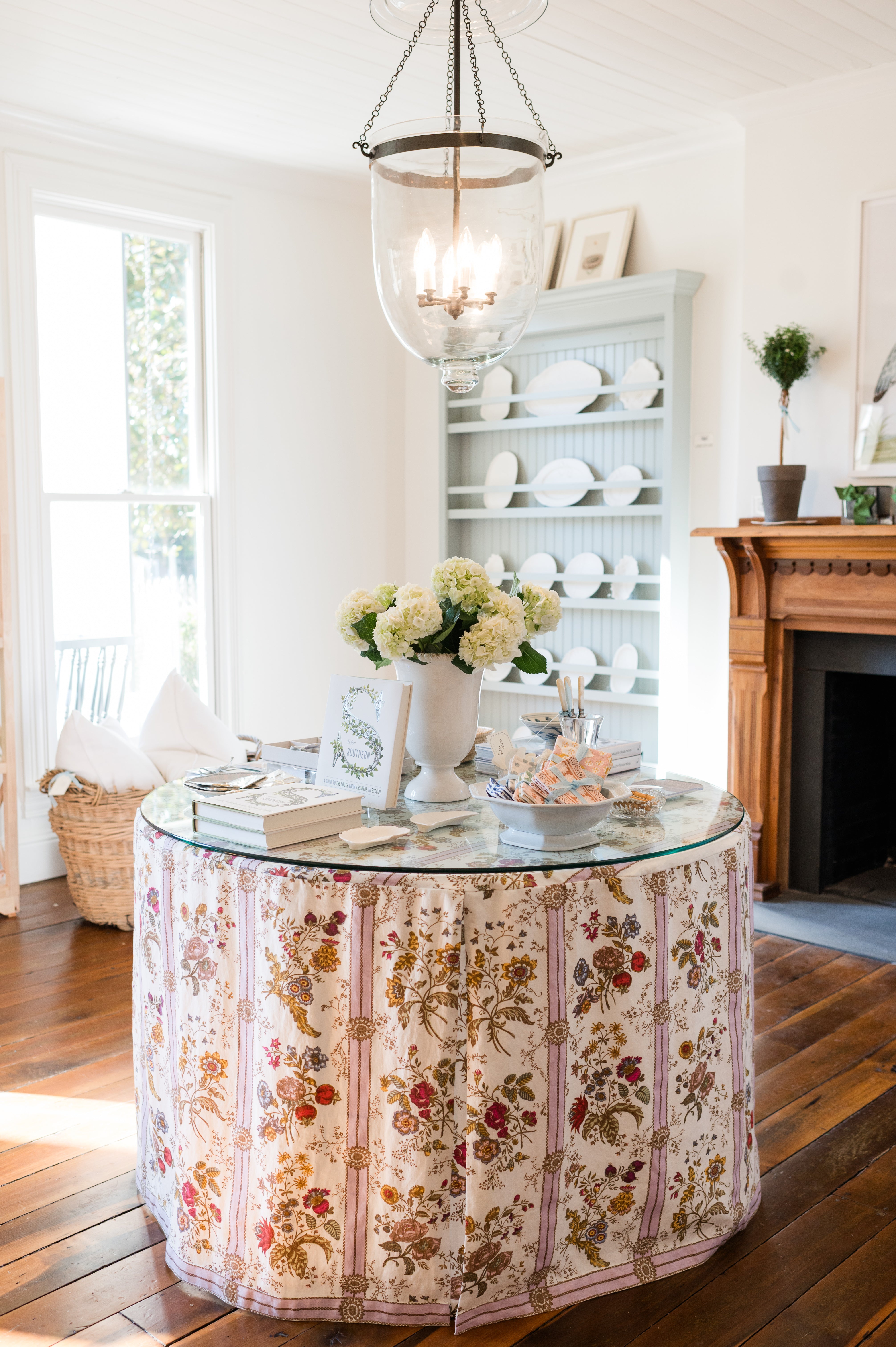 Round table with floral tablecloth in a bright room with shelves and decor.